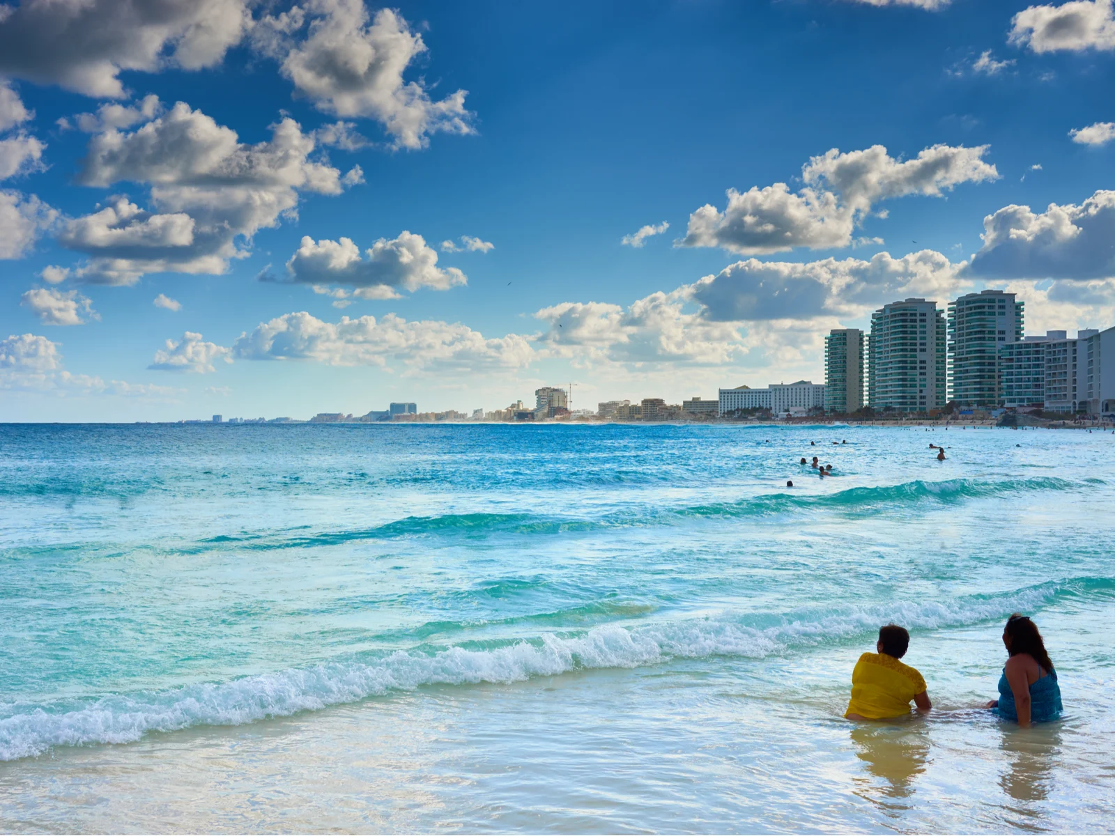 Two women dipping in the wavy shoreline while watching several people swimming at a distance in Playa Chac Mool, one of the best beaches in Cancun, near Hotel Zone