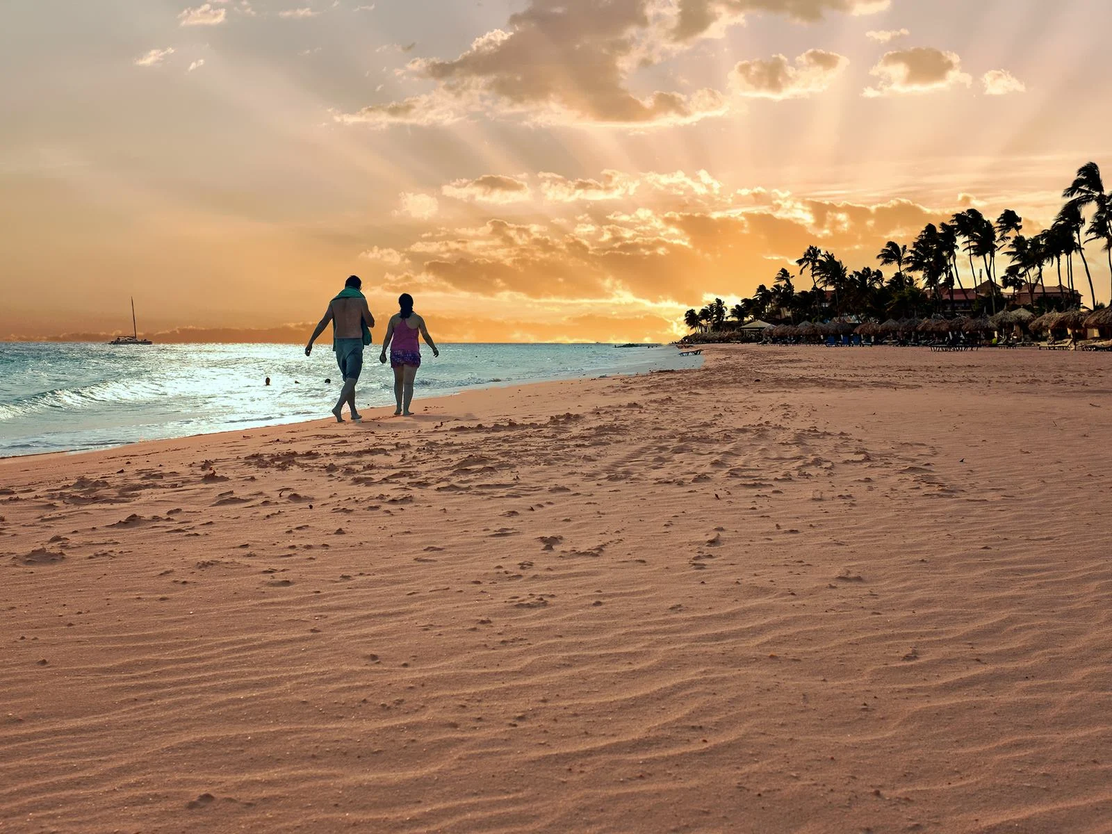 A coupe walking on the vast shoreline towards palms trees and native huts in Druif Beach during a lovely sunset with rays visible on the cloudy sky, one of the best things to do in the best beaches in Aruba