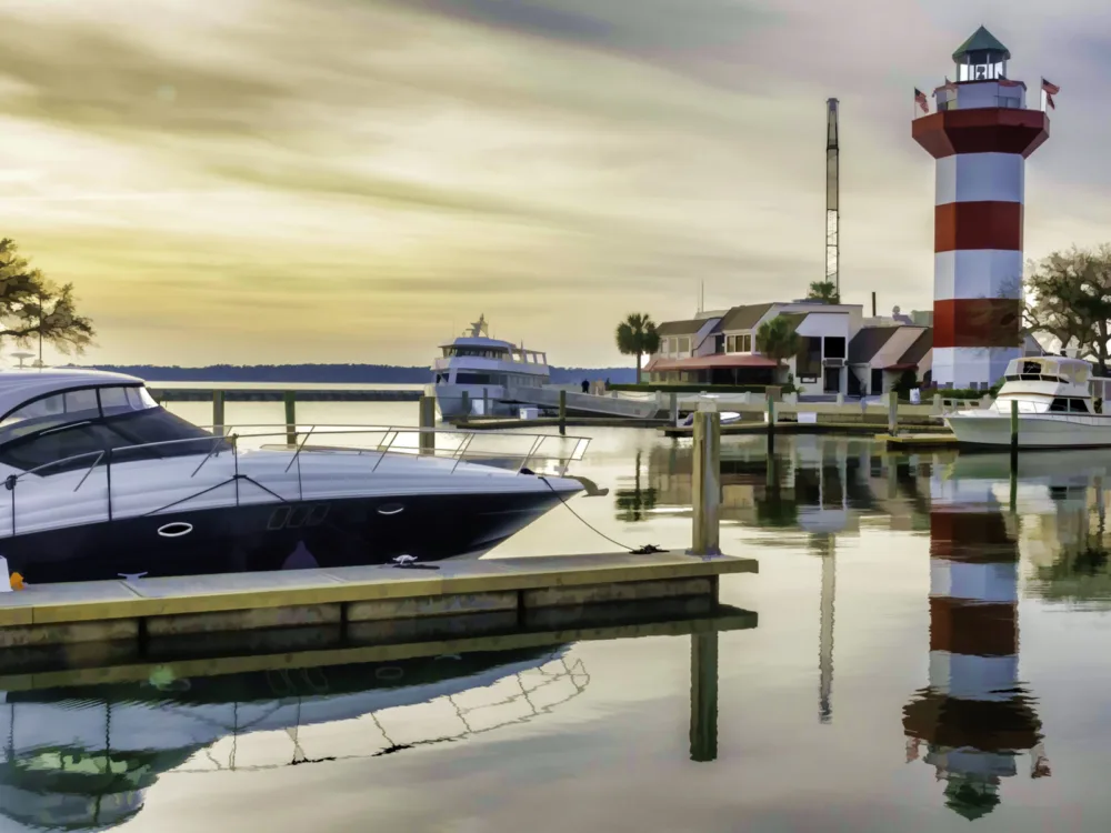 A red and white lighthouse and three docked boats reflected on the calm waters at Hilton Head Island during a spectacular late afternoon sky, titled as one of the best South Carolina attractions