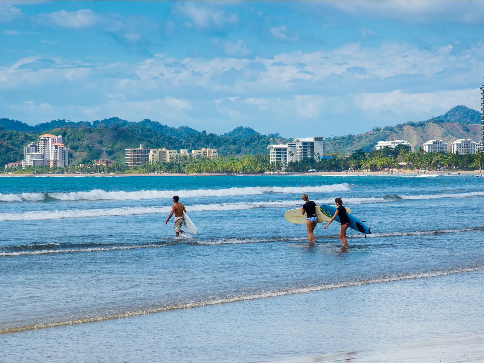 Three people carrying their surfboards towards the wavy sea of Jaco Beach, one of the best beaches in Costa Rica, during an overcast day