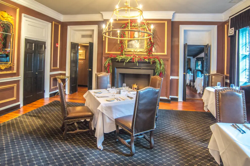 A dining table, covered with white tablecloth, and four empty chairs below a small golden chandelier in the interior of Olde Pink House, considered as one of the best tourist attractions in Georgia, and a portrait of a man hanging above the fireplace