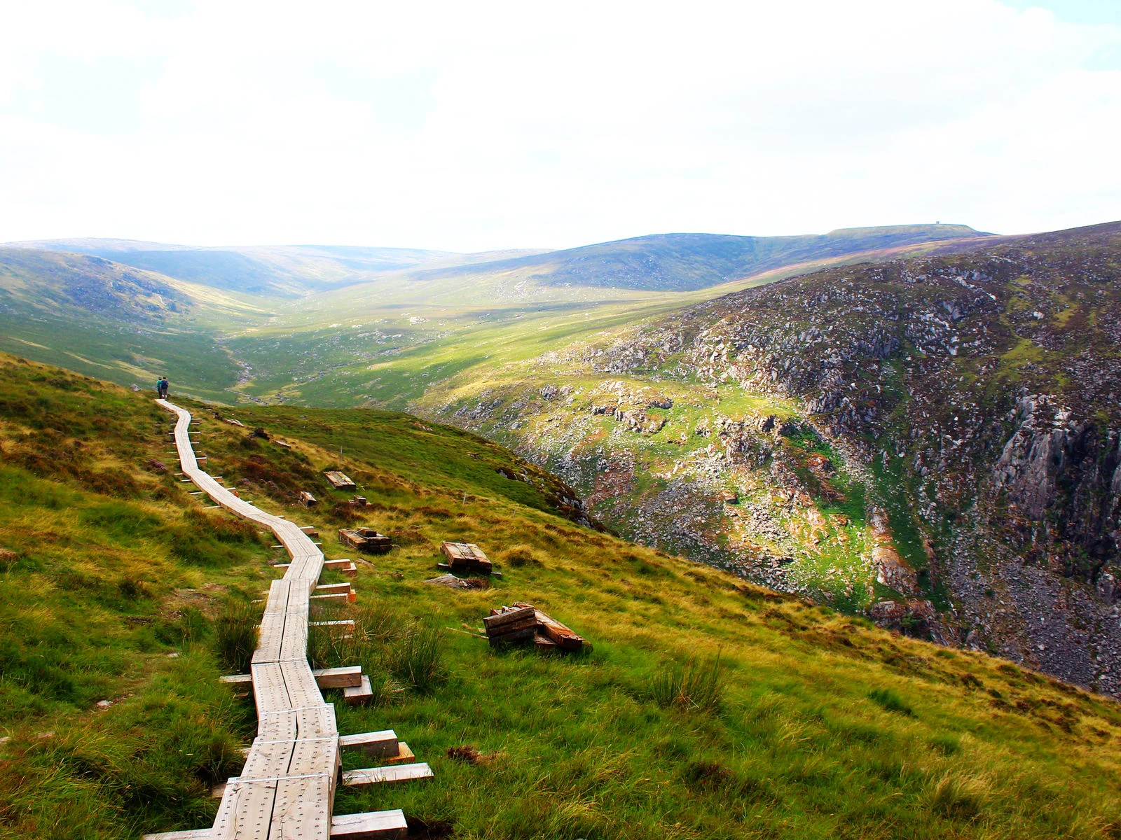 Two hikers walking on wooden planks at the scenic top of the mountain trail at Glendalough, a piece on the best hikes in Ireland, during a cold afternoon