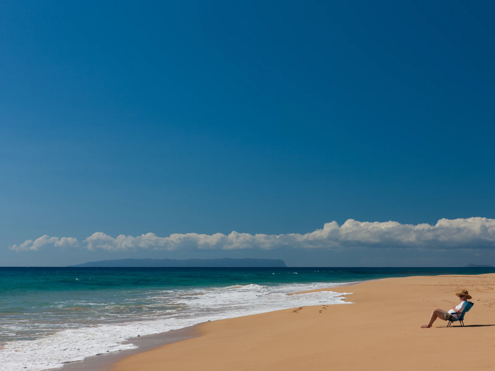 An old man wearing a sun hat sitting on a foldable chair at the golden sands of Kekaha Beach, one of the best beaches in Kauai, contemplating under the hot sun while facing the waters