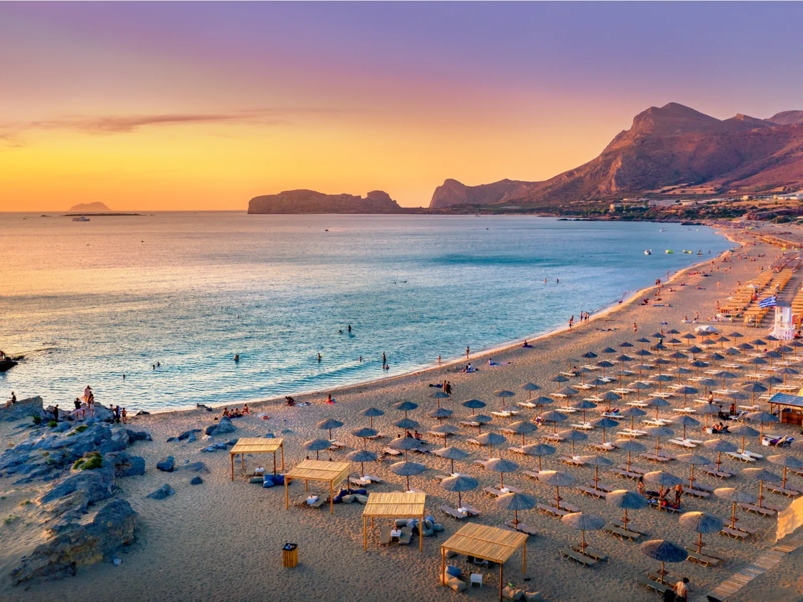 Visitors dipping and walking at the calm Falasarna Beach on a sweet sunset in Crete, one of the best islands in Greece to visit, with numerous sunbathing beds perfect arranged under beach umbrellas